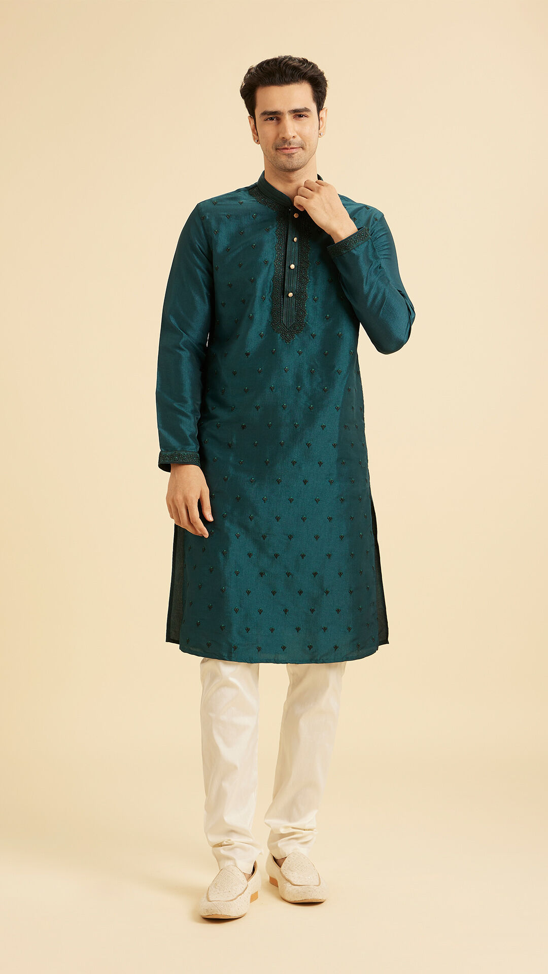 Man in a teal embroidered kurta paired with white churidar pajama, posing against a beige background in traditional Indian attire.
